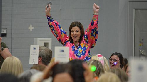 Woman standing up in a crowd of people seated at tables. Woman is celebrating victory with a joyous expression.