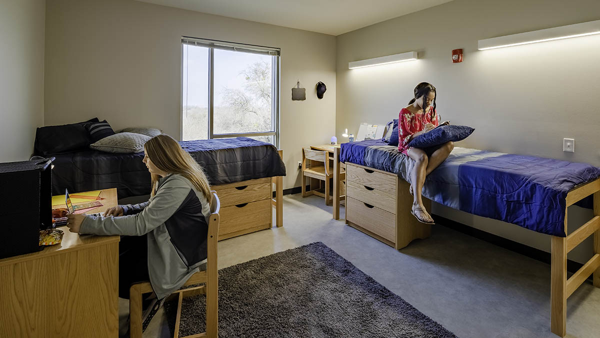 Square dorm room with one individual studying at desk and one studying on bed