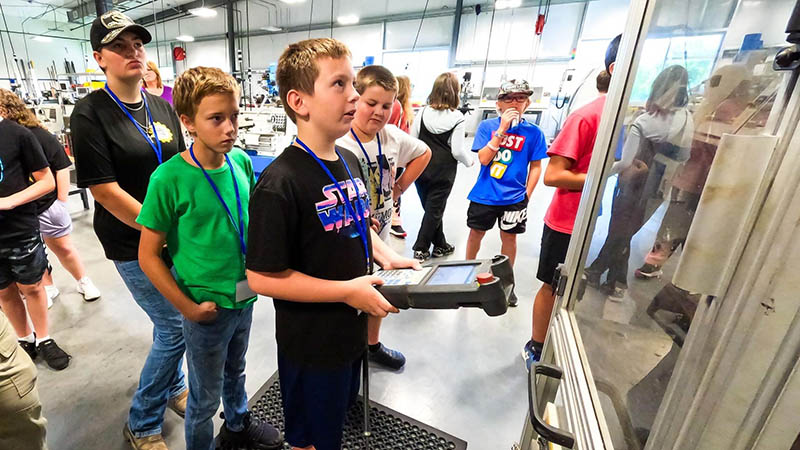 a child camper controls an industrial robot with a control panel while other camper are observing