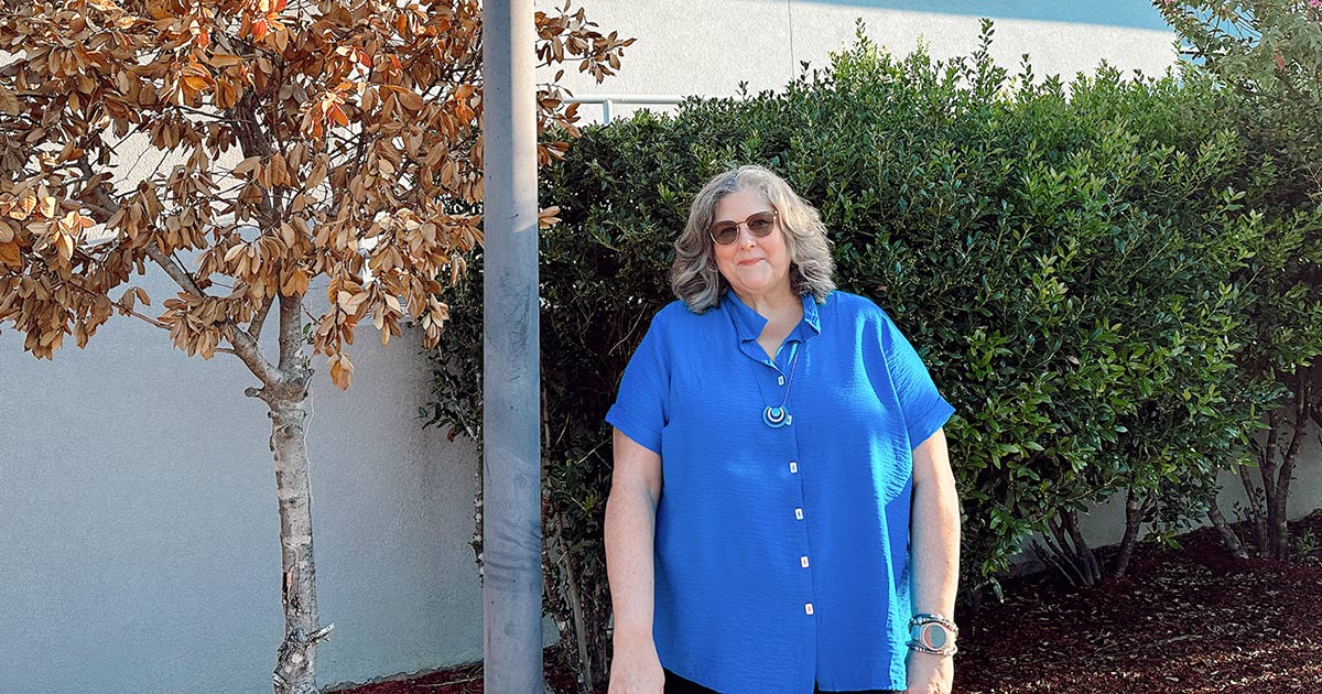 Woman wearing tinted glasses standing outside college building by bushes and tree