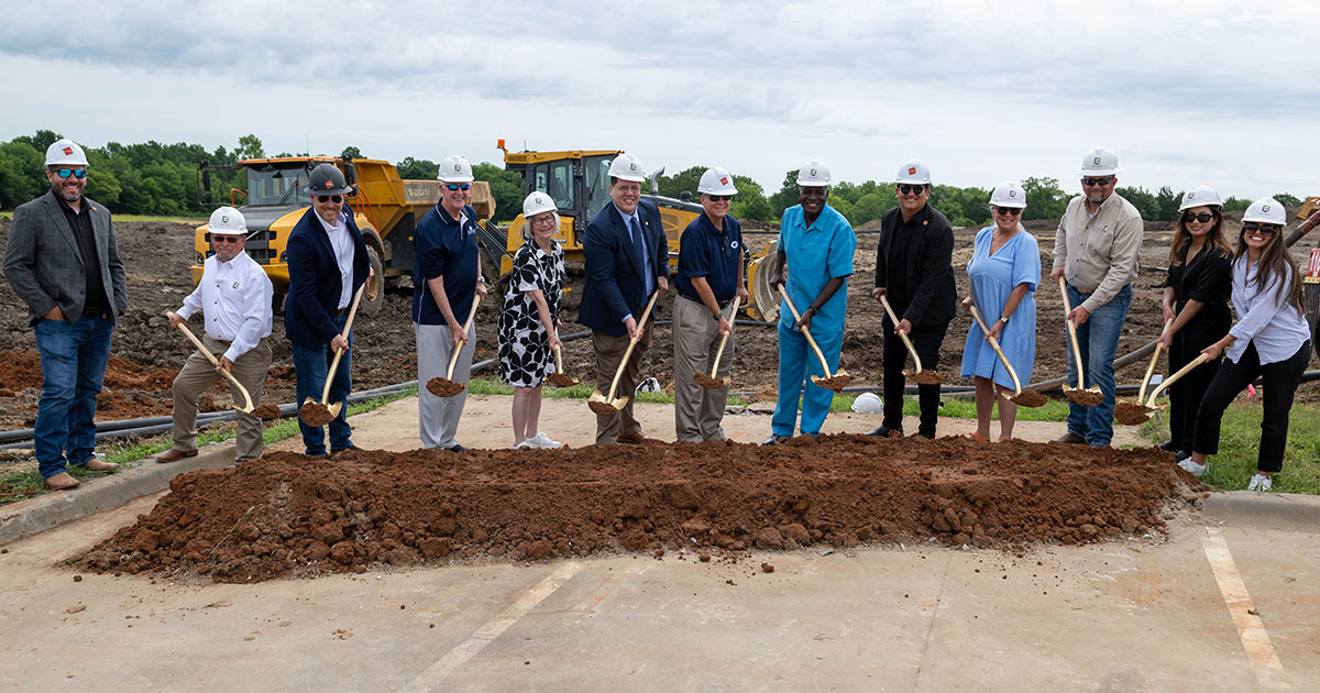 Row of people in hard hats and shovels standing in front of patch of dirt outside
